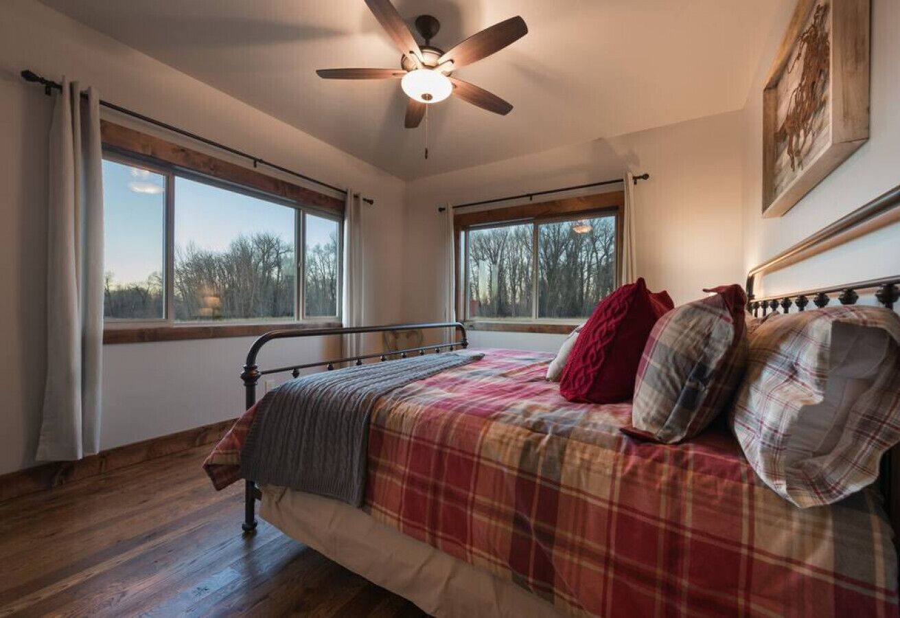 Ein Schlafzimmer mit Bett, Deckenventilator, großen Fenstern und Blick auf Bäume.