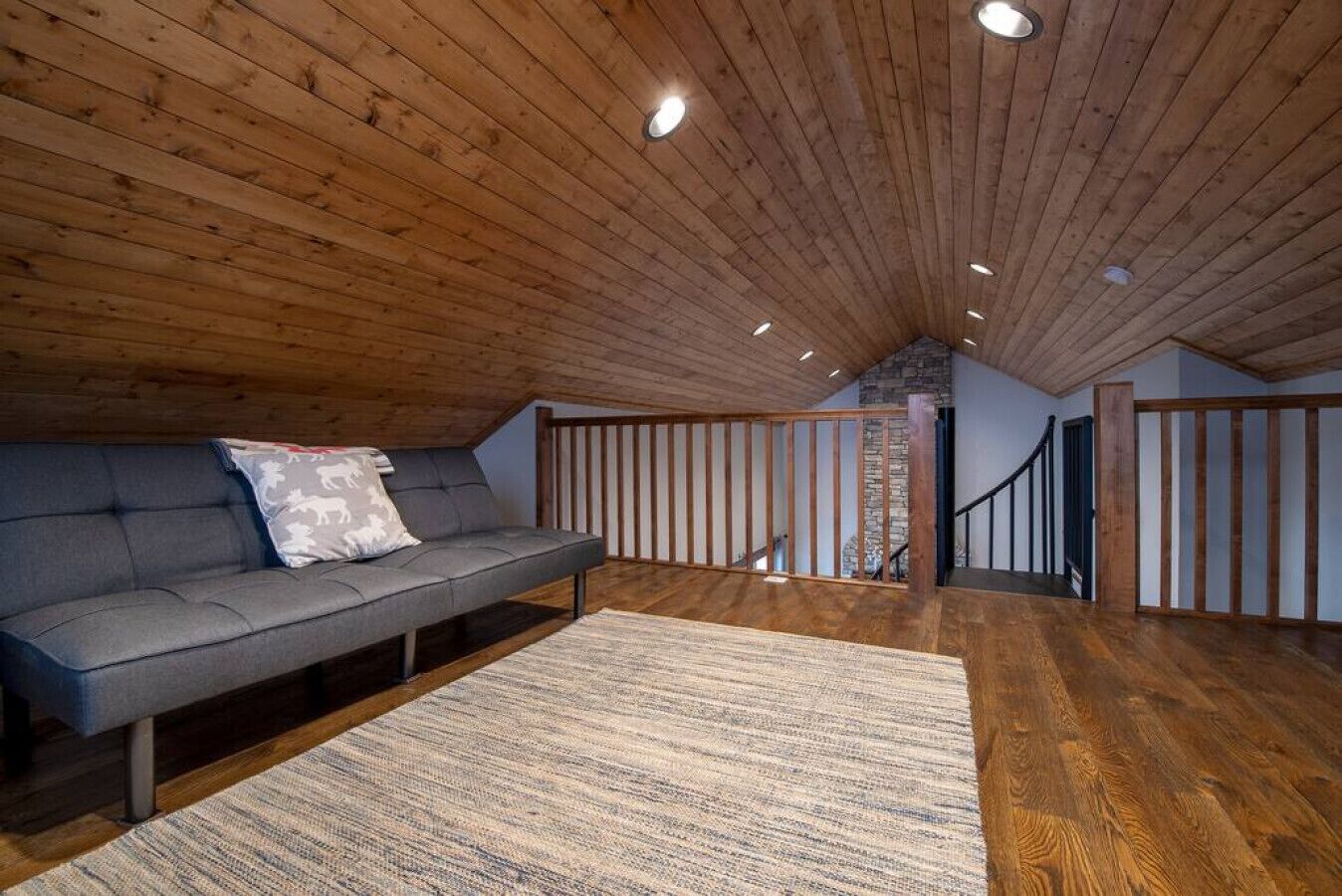 A loft-style room with a wooden ceiling, a gray sofa with patterned pillows, a woven rug, and a staircase leading to an upper level.