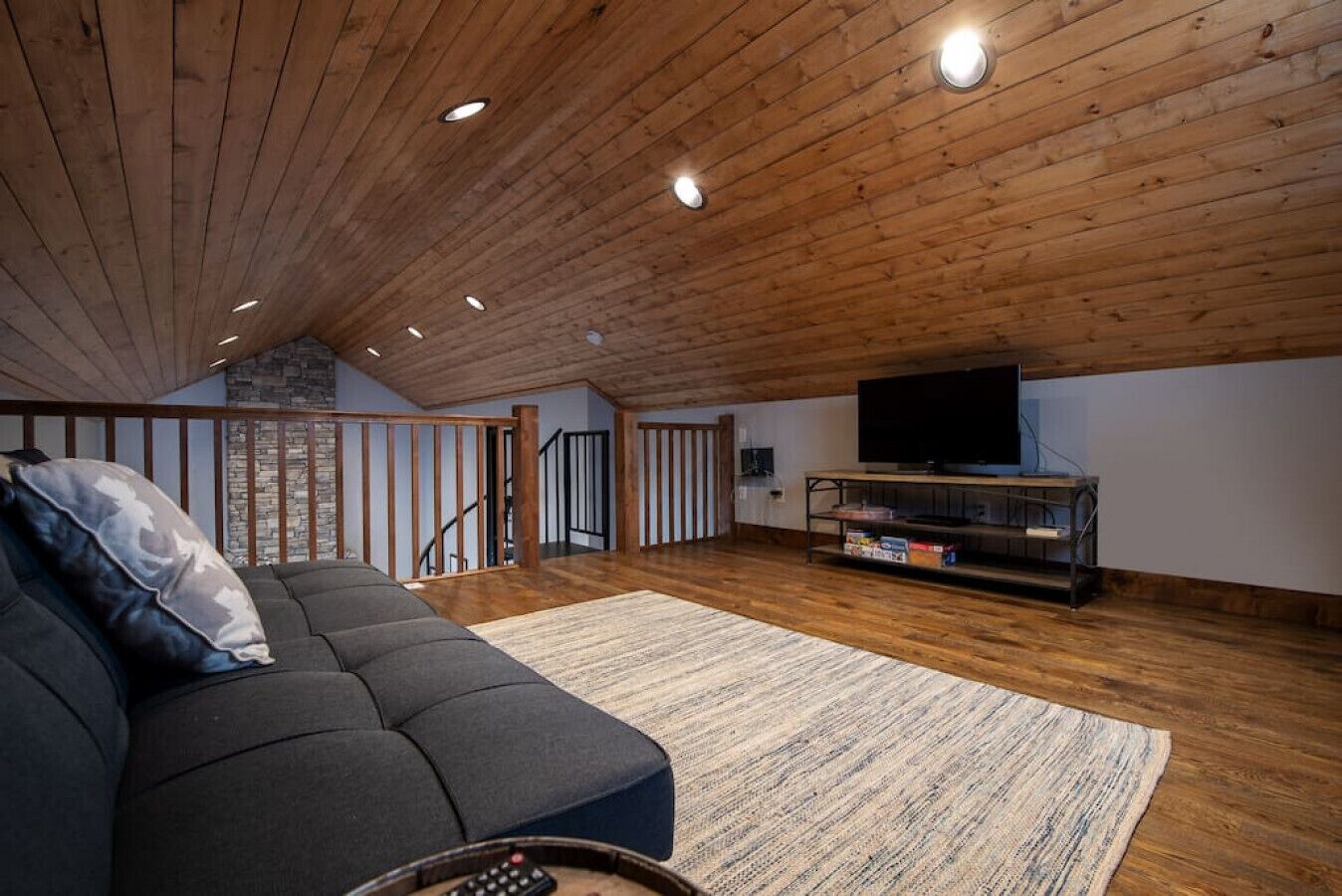 A spacious loft with a wooden ceiling, a large gray sofa, a flat-screen TV on a wooden stand, and a staircase leading to an upper level.