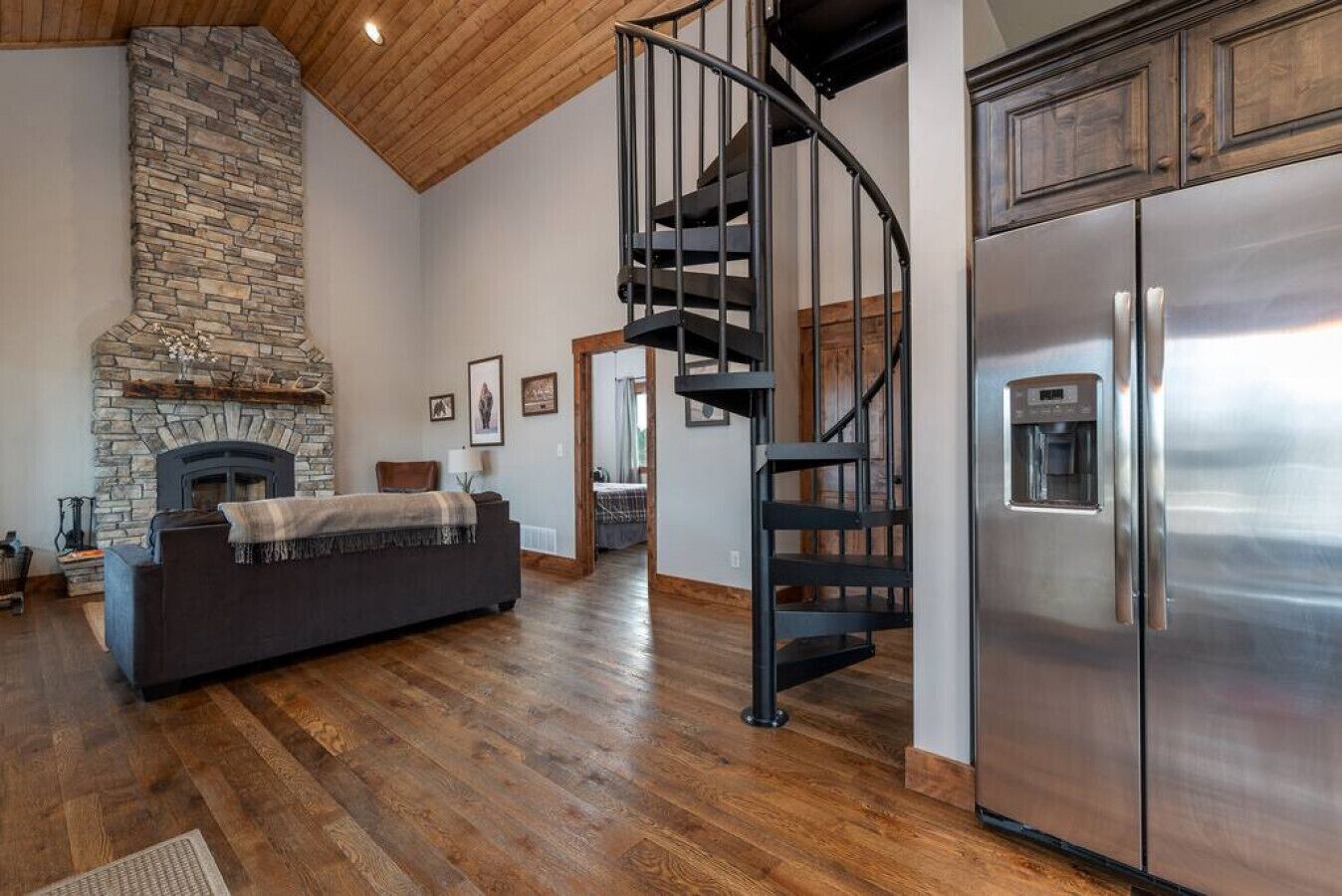 Ein modernes Wohnzimmer mit Kamin aus Stein, einem dunklen Sofa und einer Wendeltreppe, die zu einem Küchenbereich mit einem Edelstahlkühlschrank führt.