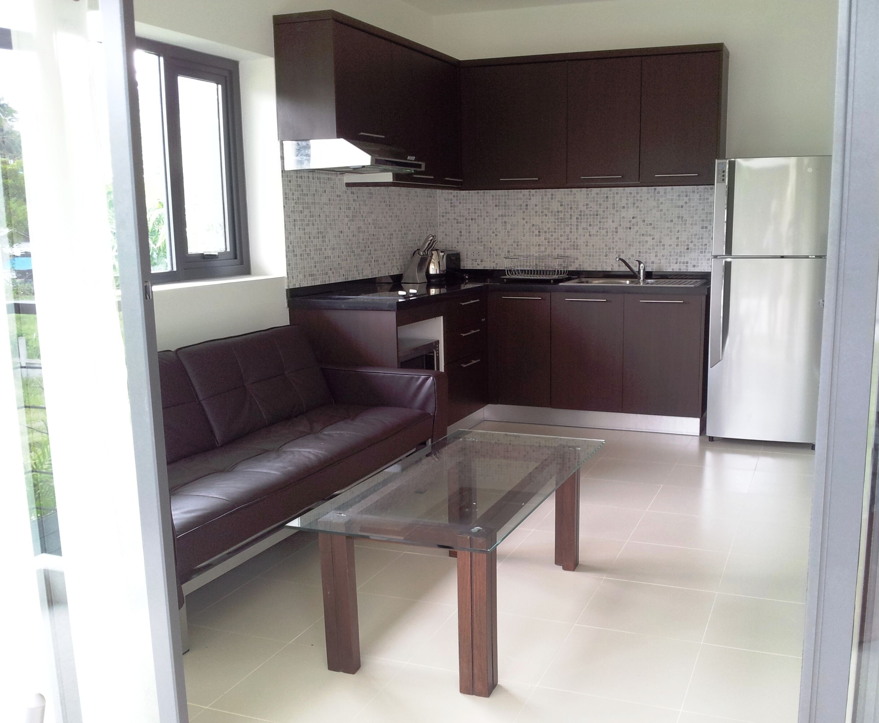A modern kitchen with dark cabinets, a glass coffee table, a sofa, and a refrigerator.
