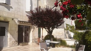 Une cour avec un sol en pierre, une table entourée de chaises et un arbre aux fleurs rouges.