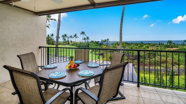 Un coin repas avec une table dressée pour six personnes, entourée de chaises, donnant sur un jardin luxuriant et l’océan.