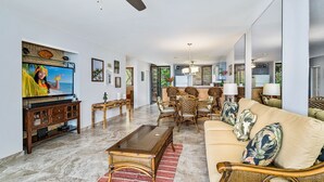 A spacious living room with a large TV, comfortable seating, and a kitchen area in the background. - 🏖️Ocean & Golf View Keauhou Condo | AC | No Stairs (Kailua- Kona)