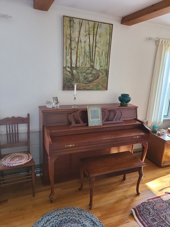 Una habitación con un piano de madera, un cuadro enmarco de un bosque, una silla de madera y una alfombra estampada.