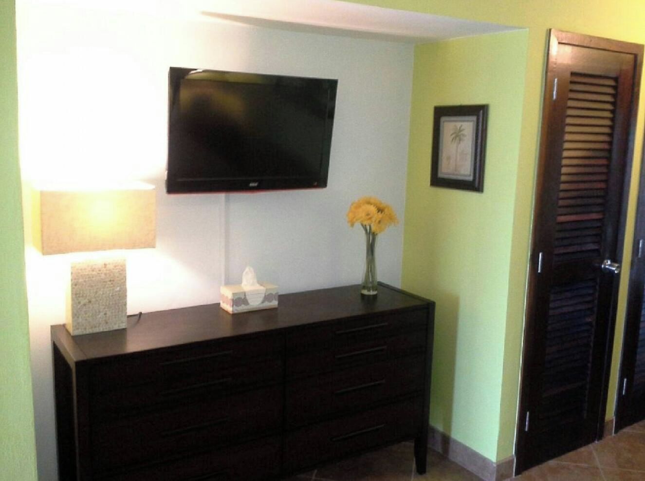 A room with a dark wooden dresser, a flat-screen TV mounted on the wall, a vase with yellow flowers, and a framed picture on the wall.
