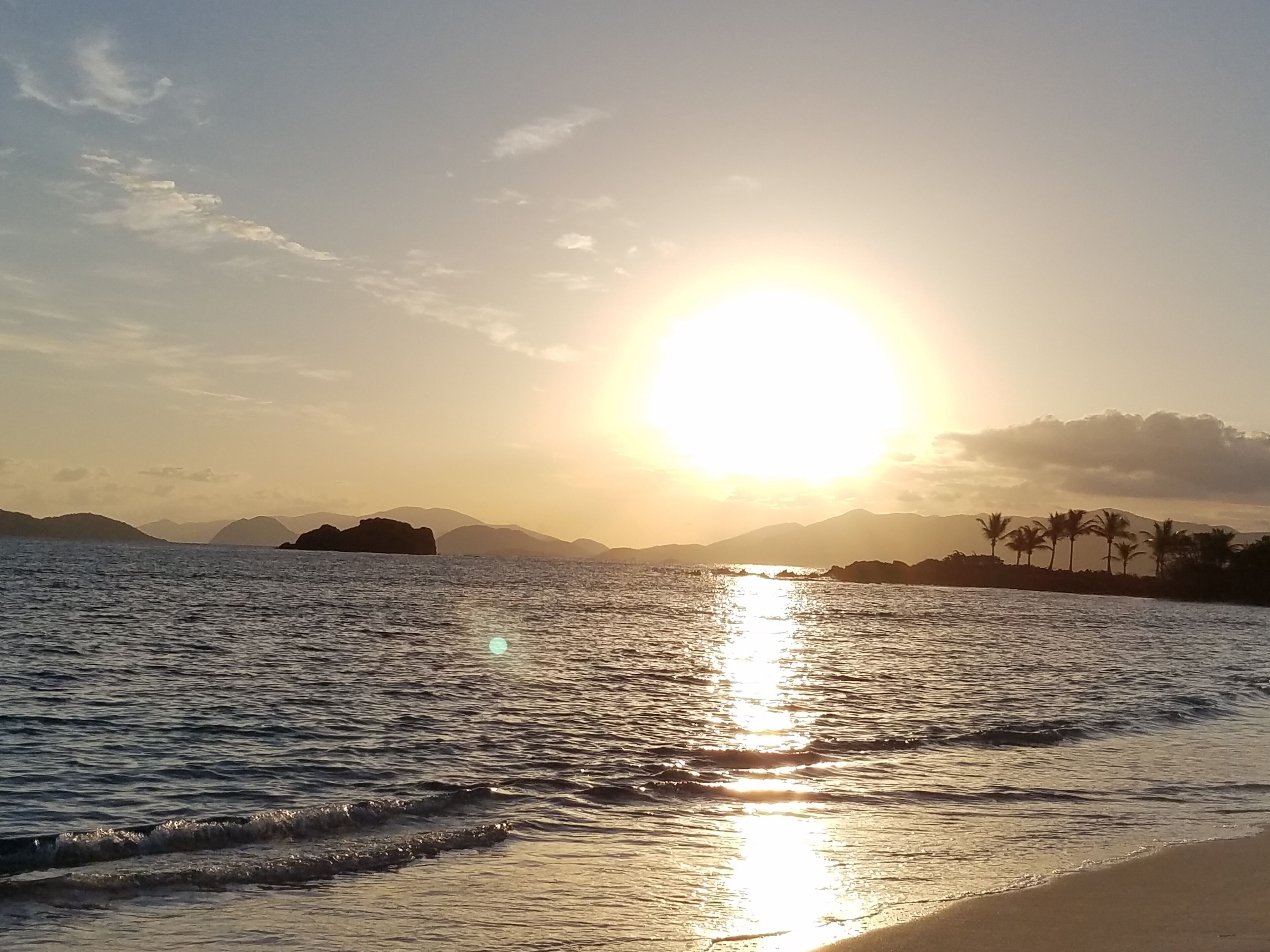 Een strand met rustig water dat de zon weerkaatst, verre bergen en palmbomen.