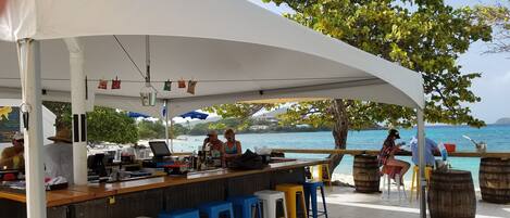 A beachside bar with a white canopy, wooden bar counter, and colorful stools.