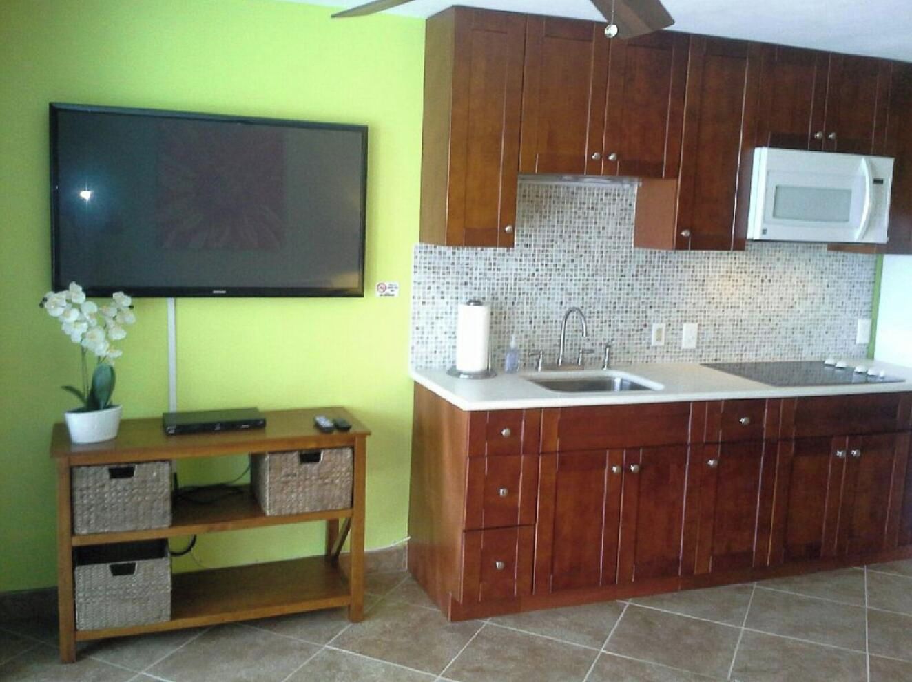 A kitchen with wooden cabinets, a microwave, a tiled backsplash, and a television on the wall.