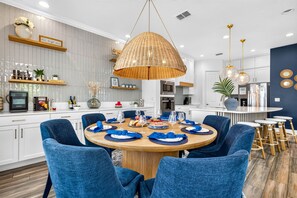 A modern kitchen with a dining area, featuring a round wooden table with blue upholstered chairs, a hanging rattan light fixture, and a built-in oven. - Tropical Treasure 4BR Villa with Swim Spa (Redington Beach)