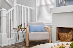 Una habitación con una escalera blanca, un sillón tapizado en azul, una pequeña mesa de madera con un adorno floral y una cesta de mimbre.