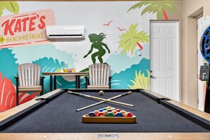 Una mesa de billar con bolas y tacos, una pared con un mural con temática playera, un letrero que dice 'KATE'S BEACH RETREAT' y una puerta con panel de seguridad.