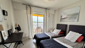 Ein Hotelzimmer mit zwei Betten, einem Schreibtisch mit Lampe, einem Sessel und Blick auf den Strand.
