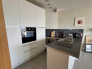A modern kitchen with white cabinets, a built-in oven, a stainless steel sink, and a small island with a cutting board.