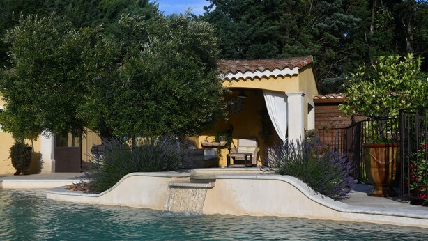 A pool with a waterfall feature, surrounded by a patio area with outdoor furniture and a small building with a tiled roof. - Gîte de provence jacuzzi (Velleron)