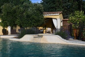 A pool with a waterfall feature, surrounded by a patio area with outdoor furniture and a small building with a tiled roof. - Gîte de provence jacuzzi (Velleron)
