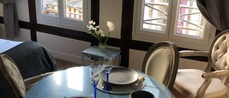 A dining area with a round glass table, two chairs, a vase with flowers, and a view of the outside through large windows.