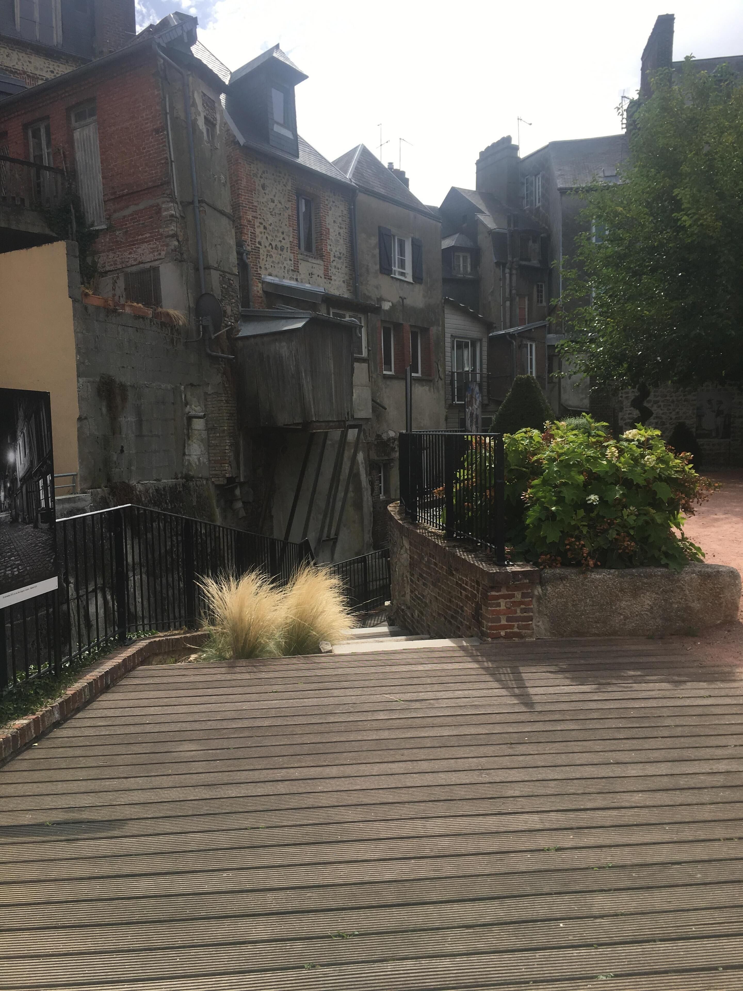 Une rue étroite bordée de vieux bâtiments, avec une promenade en bois et un petit espace aménagé en jardin.