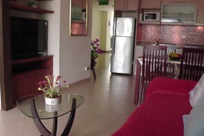 Un espace de vie compact comprenant un téléviseur, un coin repas avec une table et des chaises, une cuisine équipée d’un réfrigérateur et d’un four à micro-ondes, ainsi qu’une plante en pot.