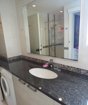 Une salle de bain avec un lavabo blanc, un grand miroir, une cabine de douche en verre et un lave-linge.