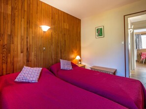 Une chambre avec deux lits, des murs lambrissés, une petite table avec une lampe et une porte donnant sur une autre pièce.