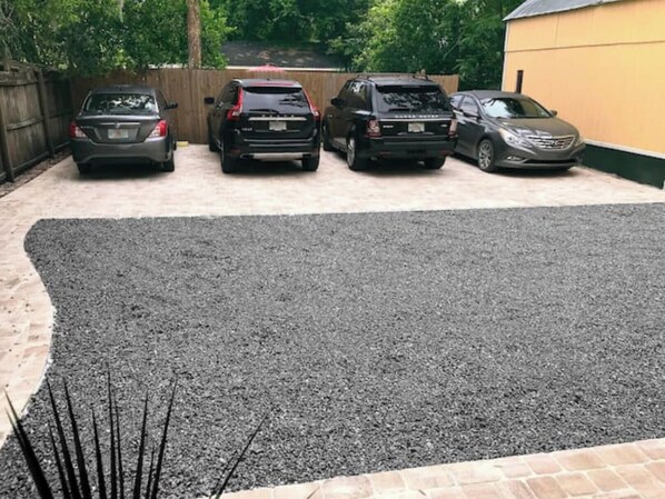 Uma área de estacionamento com cinco carros estacionados em fila, um caminho de acesso de cascalho e uma cerca de madeira ao fundo.