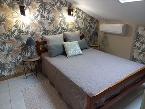 A bedroom with a bed, pillows, a bedside lamp, a small round table, a wall-mounted air conditioner, and a patterned wallpaper.
