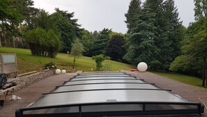 Une grande structure de toit plate et de couleur sombre, située sur une terrasse en bois, entourée d’arbres et d’une zone herbeuse.