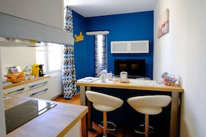Une cuisine compacte avec un mur d’accent bleu, une machine à café, deux tabourets de bar et un téléviseur.