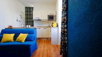 A compact living space with a blue sofa, a kitchenette with a microwave, and a wooden floor.