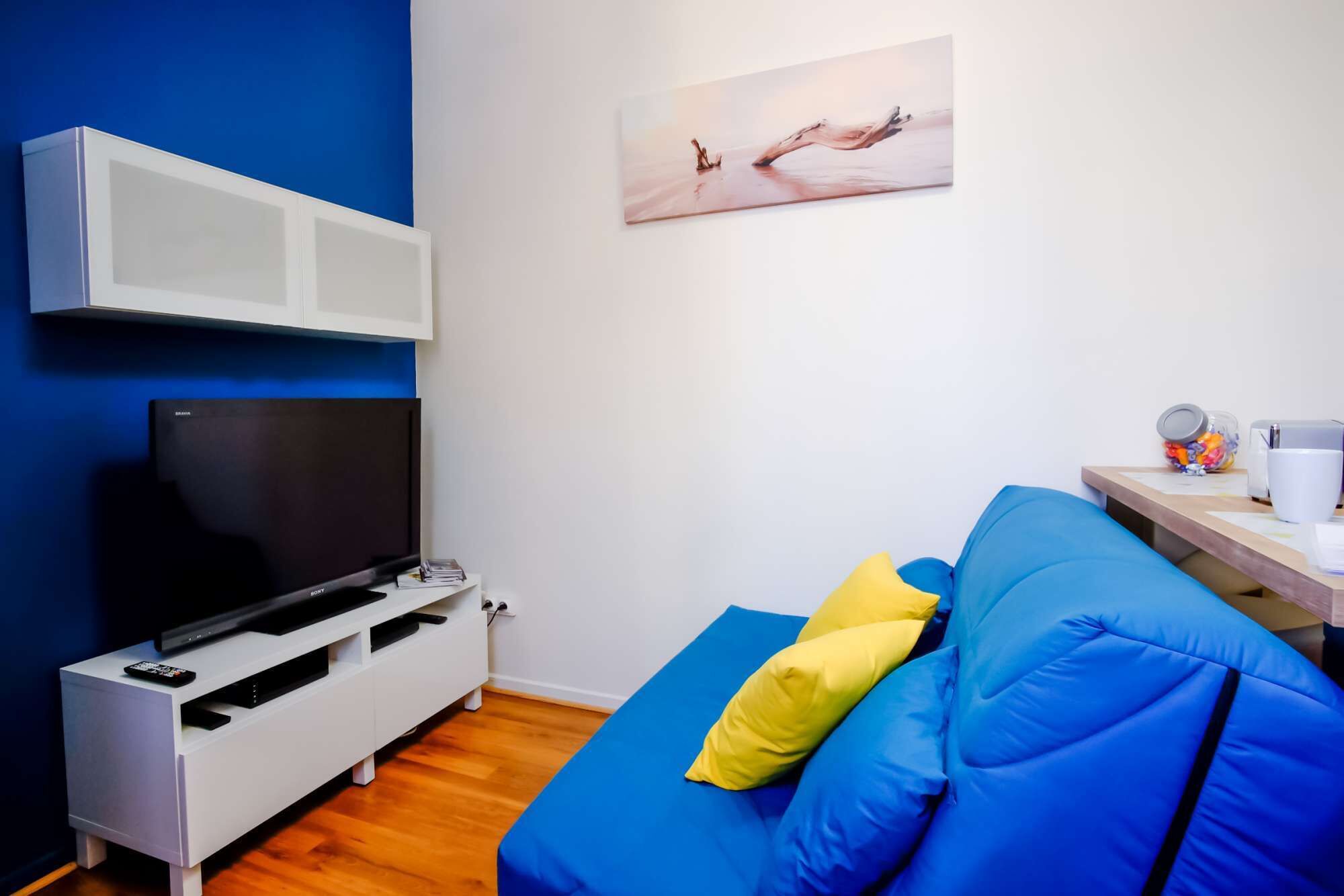 Un salon moderne avec un canapé bleu, un téléviseur à écran plat, un meuble blanc et un tableau accroché au mur.