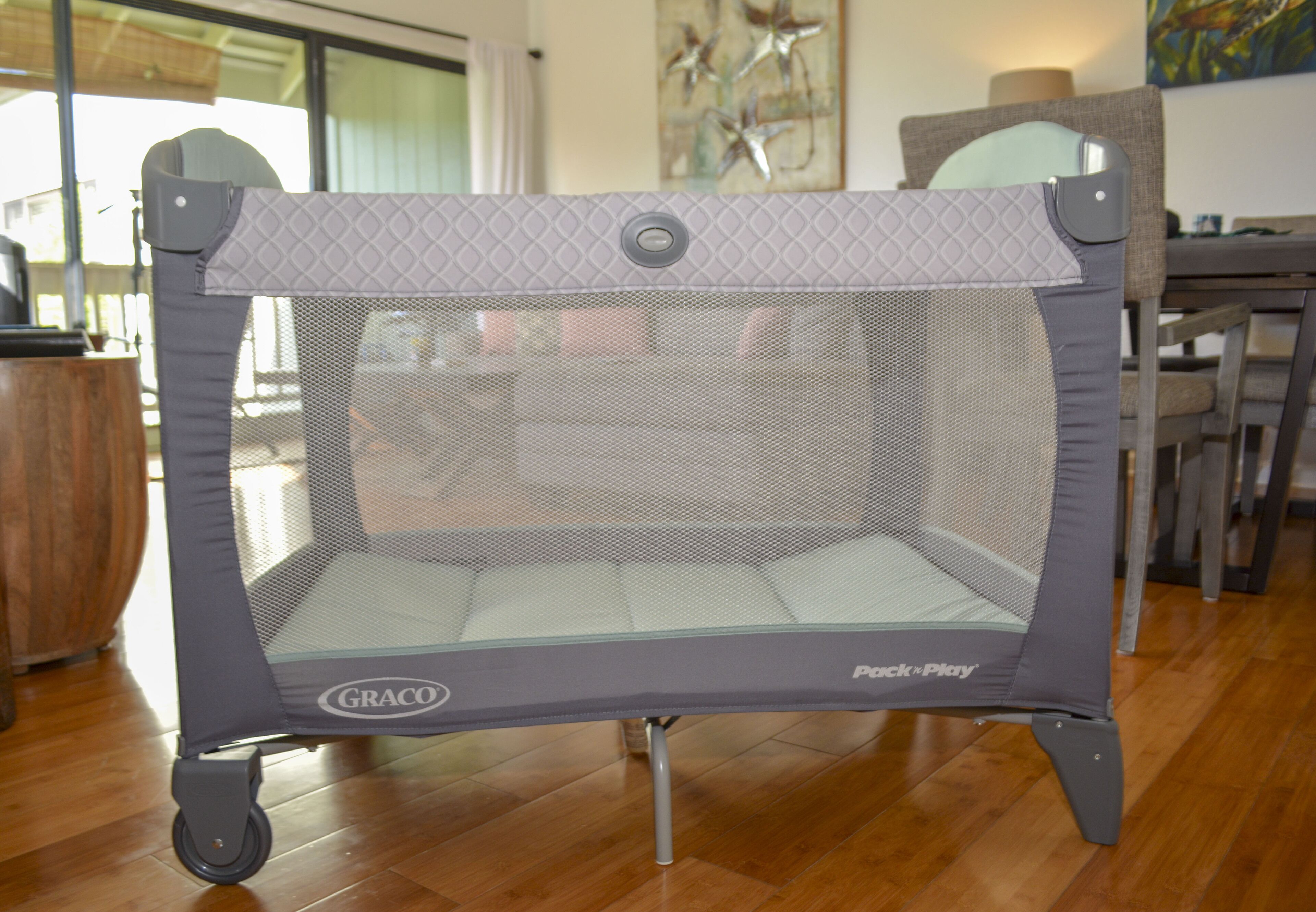 Een Graco Pack 'n Play reisbed met wielen, geplaatst op een houten vloer in een kamer met een eethoek op de achtergrond.