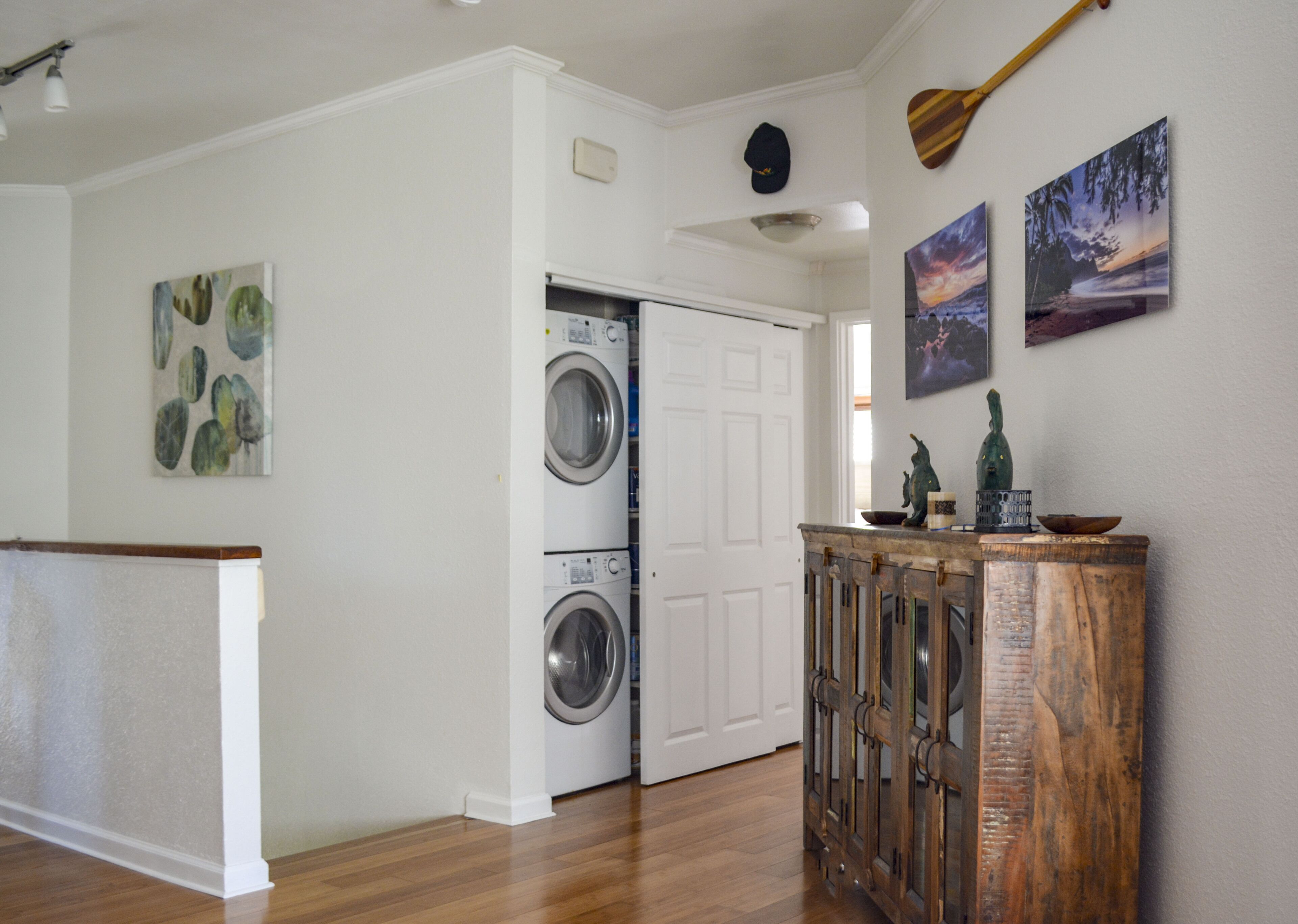 A laundry room with two stacked front-loading washers and dryers, a wooden cabinet with decorative items, and a door leading to another room.