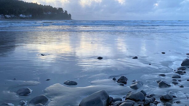 A beach with rocks and calm water, a cloudy sky, and a forested hill in the background.