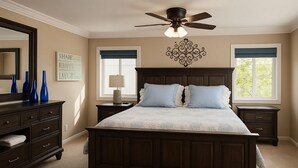 Un dormitorio con una cama grande, cabecera de madera oscura, dos mesitas de noche, una mesita auxiliar, un ventilador de techo y una ventana con vistas a árboles.