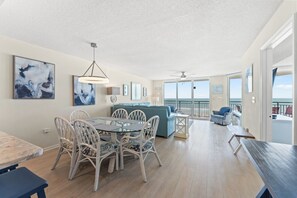 A spacious living area with a dining table, chairs, and a sofa. There is a balcony with a view of the ocean.