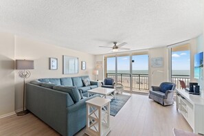 Un salón amplio con un sofá grande, una mesa de centro y una televisión. Hay un balcón con vistas a la playa.