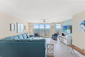 A living room with a sofa, chairs, a TV, and a balcony with a view of the ocean.
