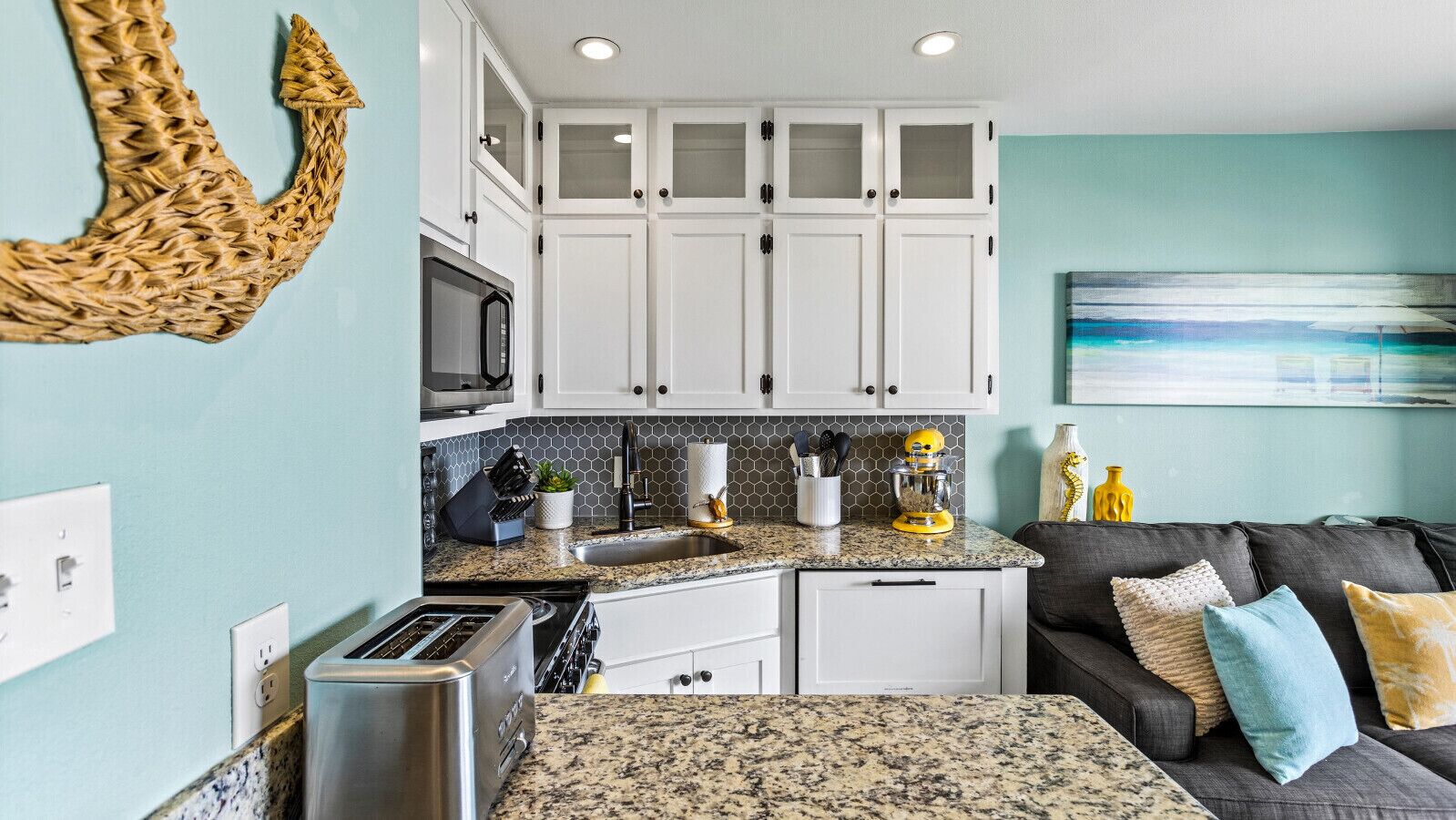 A compact kitchen with white cabinets, a microwave, a toaster, and a sink. There's a black couch with cushions and a painting on the wall.