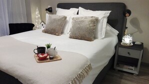 Una cama bien hecha con ropa blanca, dos almohadas, una mesita de noche con una lámpara y una bandeja con una taza, un bol y una pequeña planta.