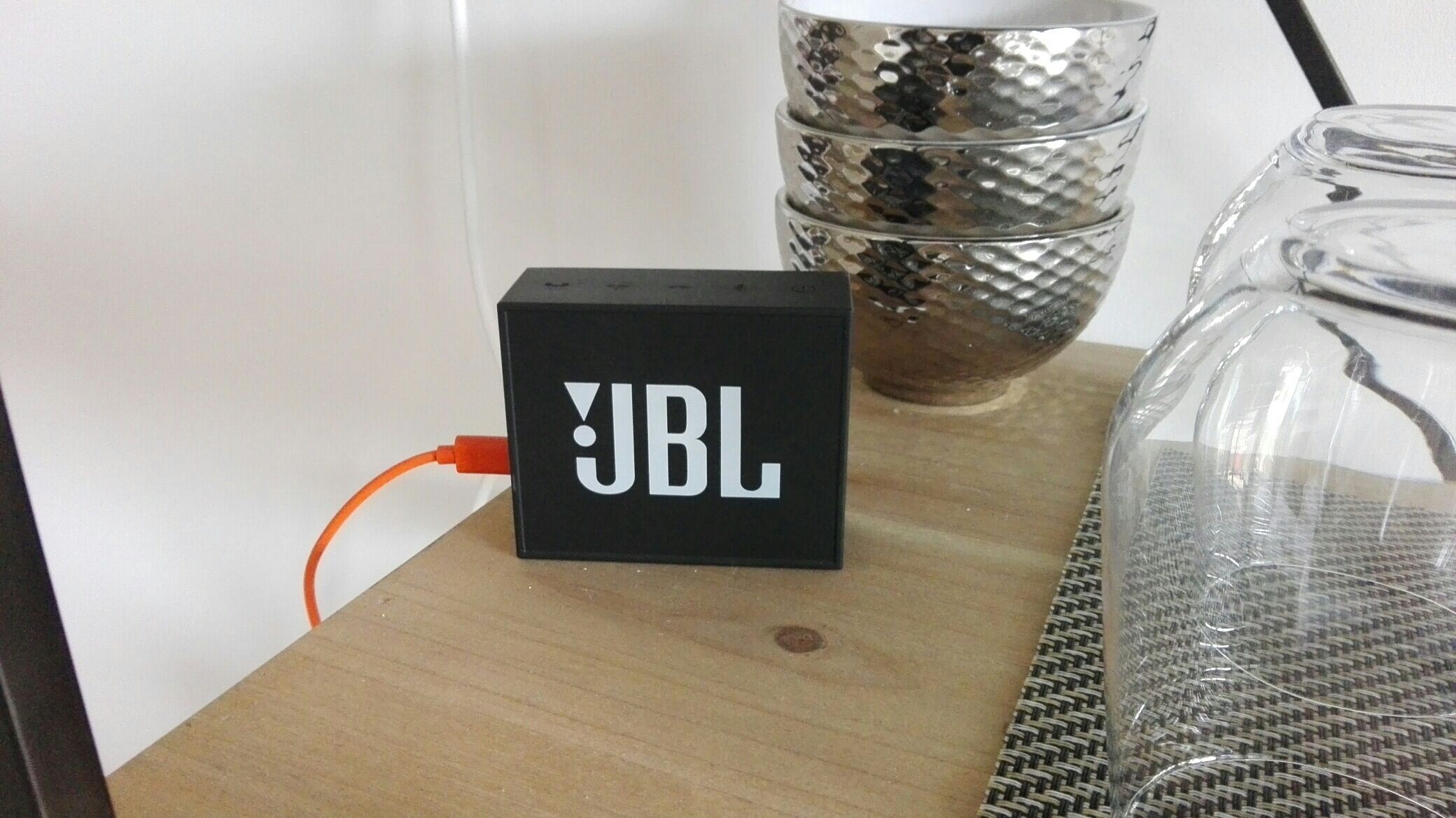 A black JBL speaker on a wooden surface with a woven mat beside it.
