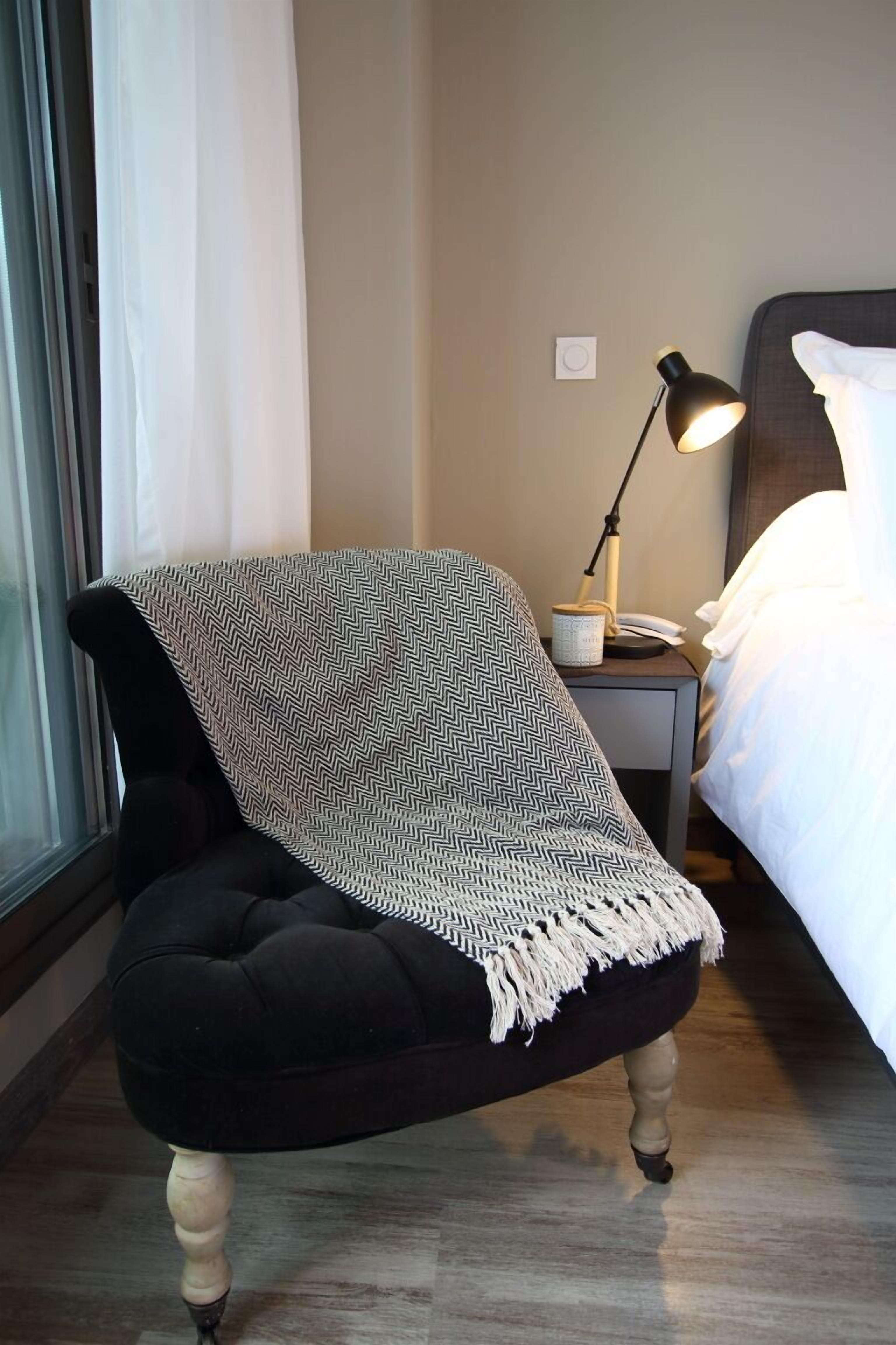 Una poltrona nera imbottita con una coperta a motivi, un comodino con una lampada e un letto con biancheria bianca.