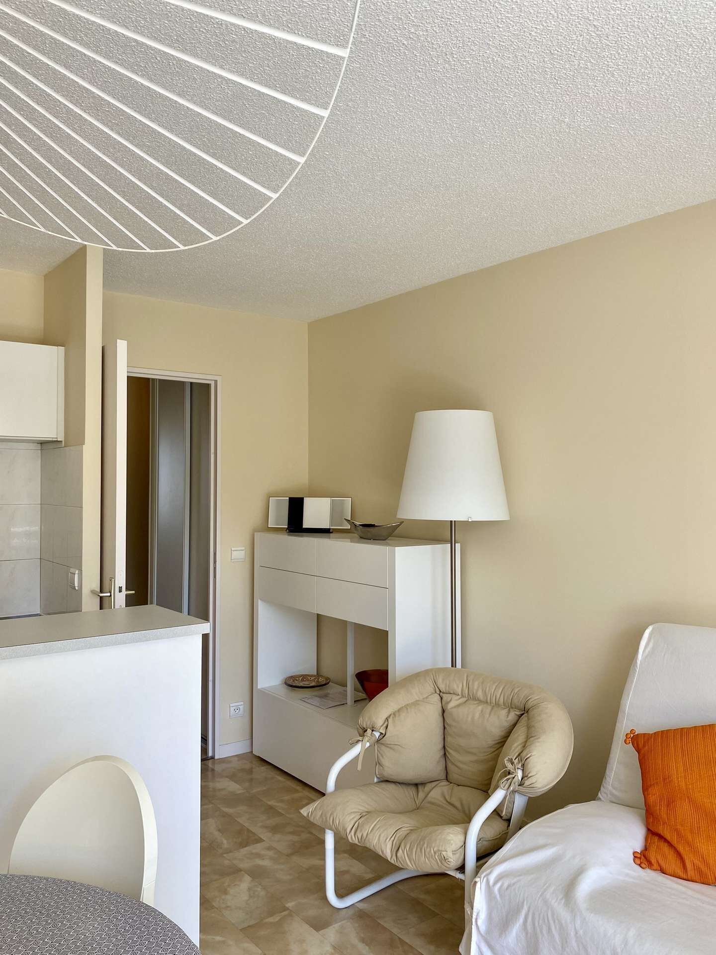 A modern hotel room with a white desk, a chair, a bed with an orange pillow, and a lamp.