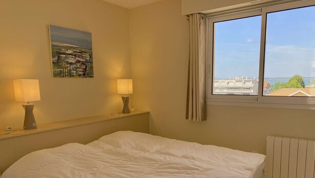 Quarto de hotel com uma cama, duas luminárias de cabeceira, um quadro na parede, uma janela com vista para edifícios e vegetação, e um radiador.