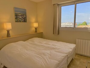 A hotel room with a bed, two bedside lamps, a painting on the wall, a window with a view of buildings and greenery, and a radiator. - Charmant T2 à Arcachon, à deux pas du centre-ville (ARCACHON)