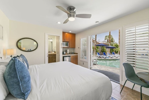 A hotel room with a bed, a ceiling fan, a bathroom with a mirror, a kitchenette, and a view of a pool area.