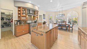 A spacious kitchen with wooden cabinets, a central island, and a dining area with a view. - California Paradise: #231868 3BR Pool, Spa, BBQ! (La Quinta)