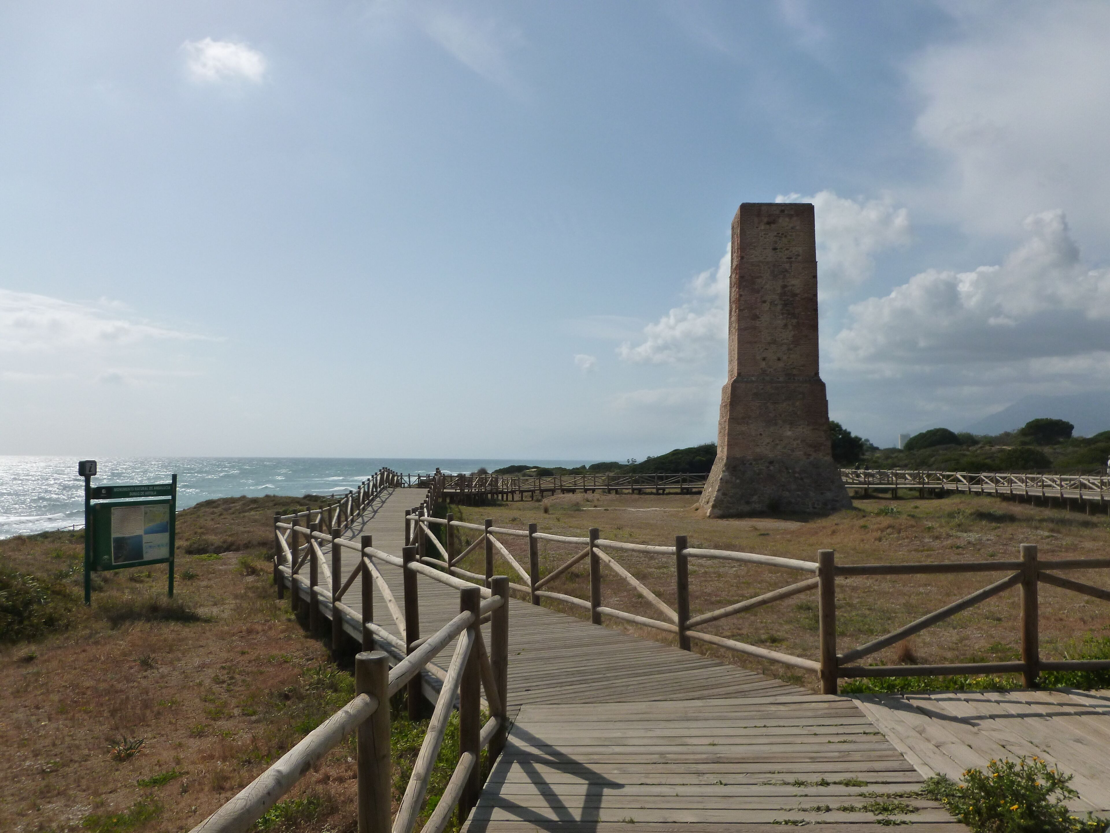 En strandpromenad som leder till ett högt stenmonument, med en träspång och en informationsskylt.