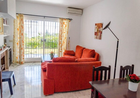 Una sala de estar con un sofá rojo, una mesa de comedor con sillas, una planta en maceta y vistas a través de un ventanal con cortinas.