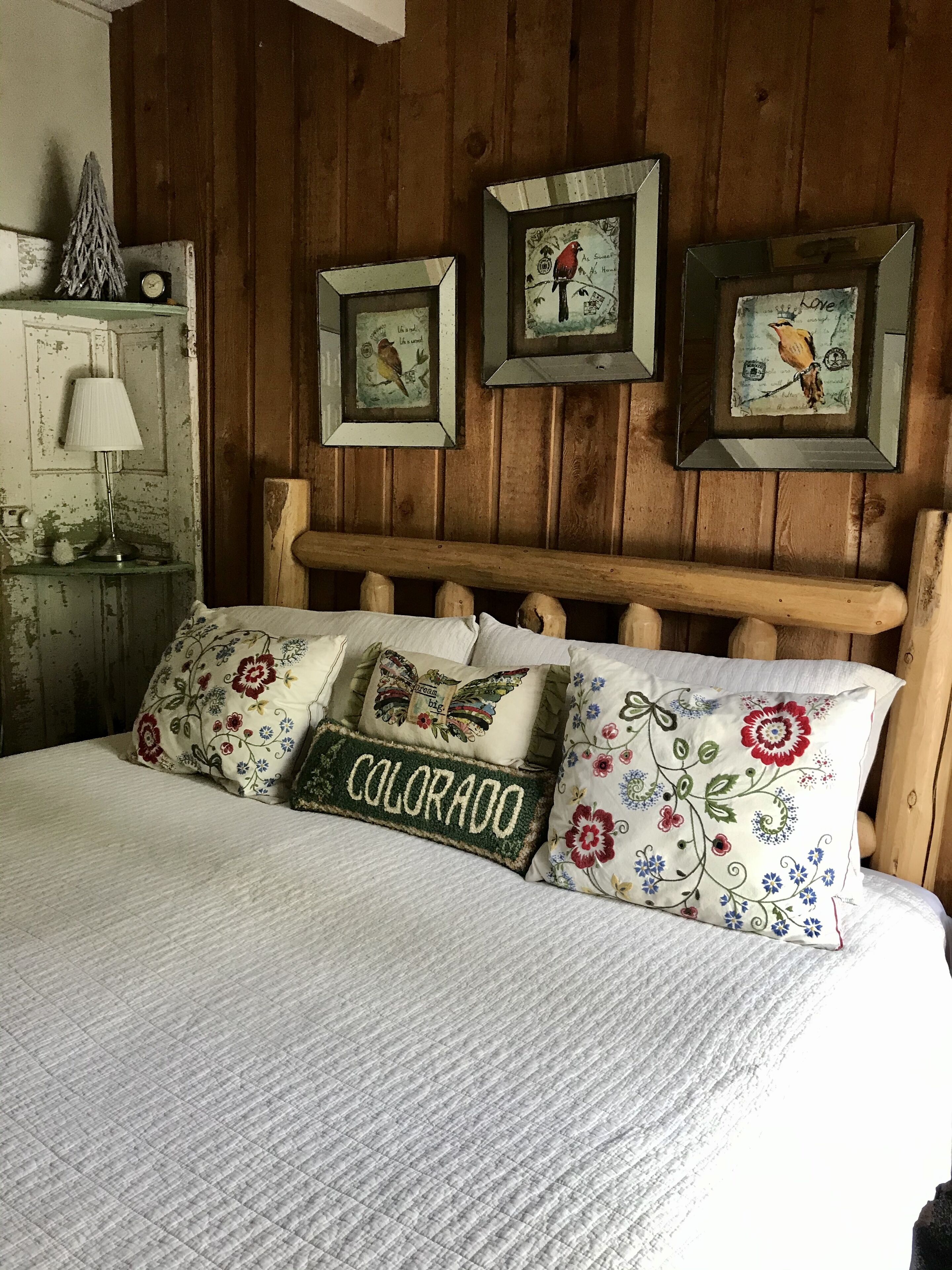 Una cama con almohadas de motivos florales y de pájaros, un cabecero de madera y decoración rústica en la pared.